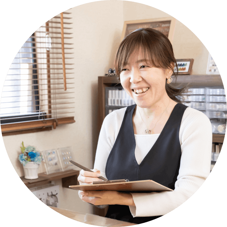 Smiling woman in a navy apron writes on a clipboard in a cozy shop or cafe setting.